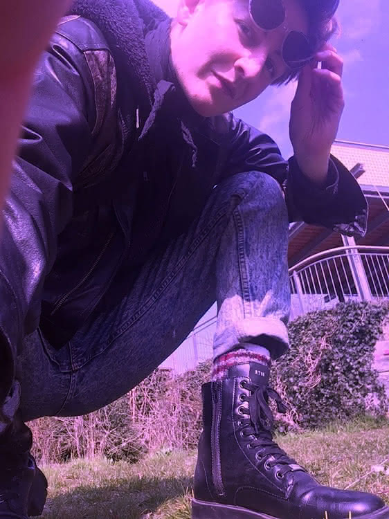 A person with ankle boots squatting close to the camera. They are holding up shades on their forehead and looking into the camera.