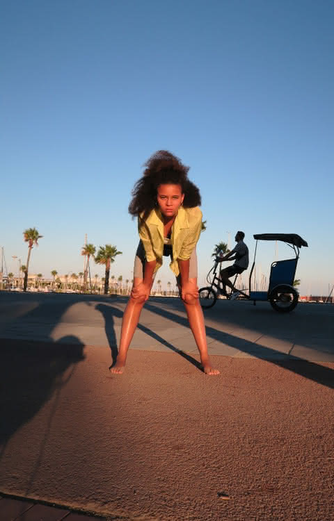 A person standing that rests their hands on their knees, looking at the camera. There are palm trees in the background.