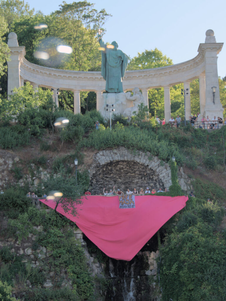 Sicht von Unten auf eine Klippe. Auf der oberen Klippe ist ein Denkmal mit einer Statue. Darunter halten Menschen einen großen rosa Winkel als Banner und darüber einen "Free Maja"-Banner.